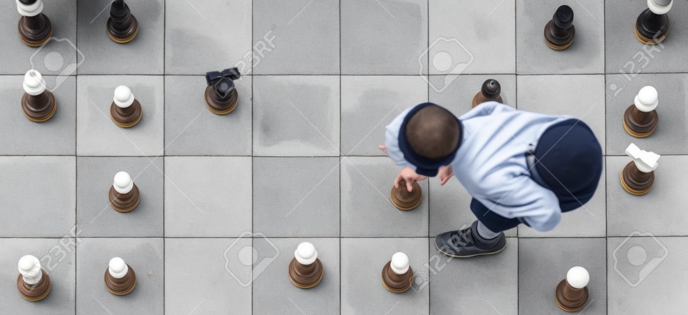 从上面看到的户外棋盘上的象棋游戏中，一个小男孩在移动一个白色棋子