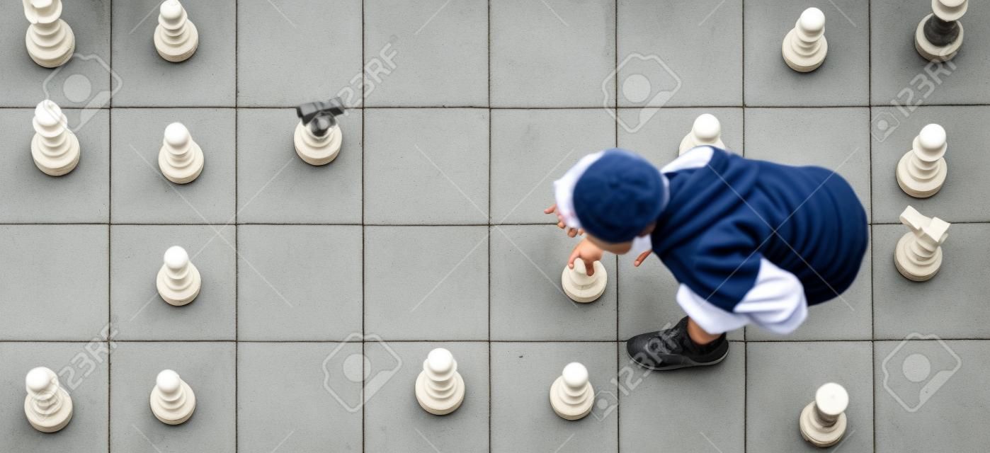 从上面看到的户外棋盘上的象棋游戏中，一个小男孩在移动一个白色棋子
