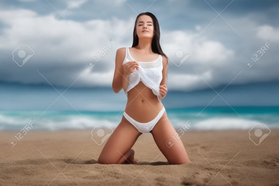 Aantrekkelijk jong vrouwtje in de top en slipje sluiten ogen en aanraken van haar terwijl staan op knieën op zandstrand tegen bewolkte hemel op het resort