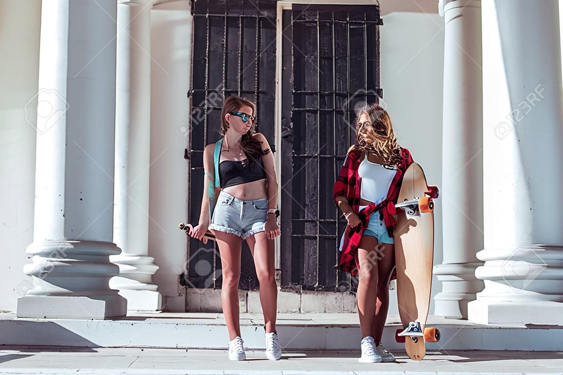 Two girls girlfriends summer city, fashionable students skateboard, longboard casual wear. Holiday weekend sunny day. Communication in street. Against background white columns large metal door.