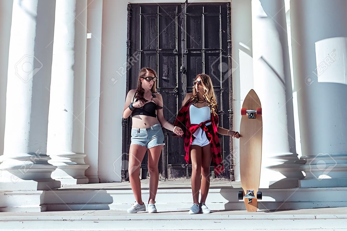 Two girls girlfriends summer city, fashionable students skateboard, longboard casual wear. Holiday weekend sunny day. Communication in street. Against background white columns large metal door.