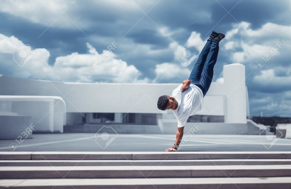 young stylish man standing one arm, guy dancer, summer city, dancing break dance, healthy fitness athletes life style, white t-shirt jeans. Dancing to music. Performs in public. Free space for text