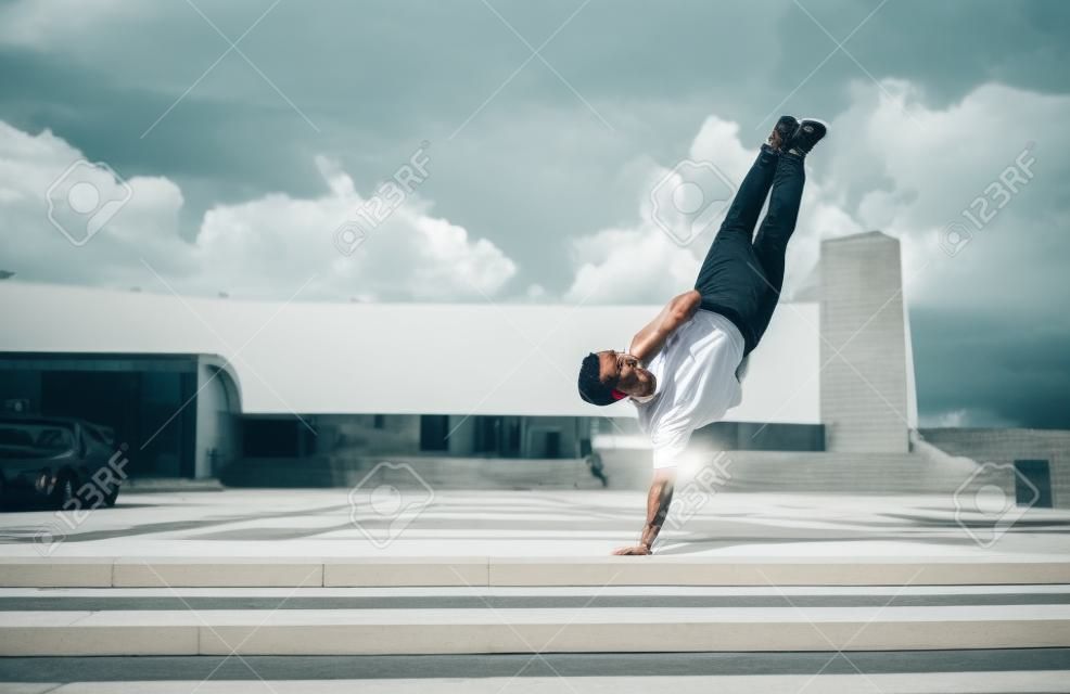 young stylish man standing one arm, guy dancer, summer city, dancing break dance, healthy fitness athletes life style, white t-shirt jeans. Dancing to music. Performs in public. Free space for text