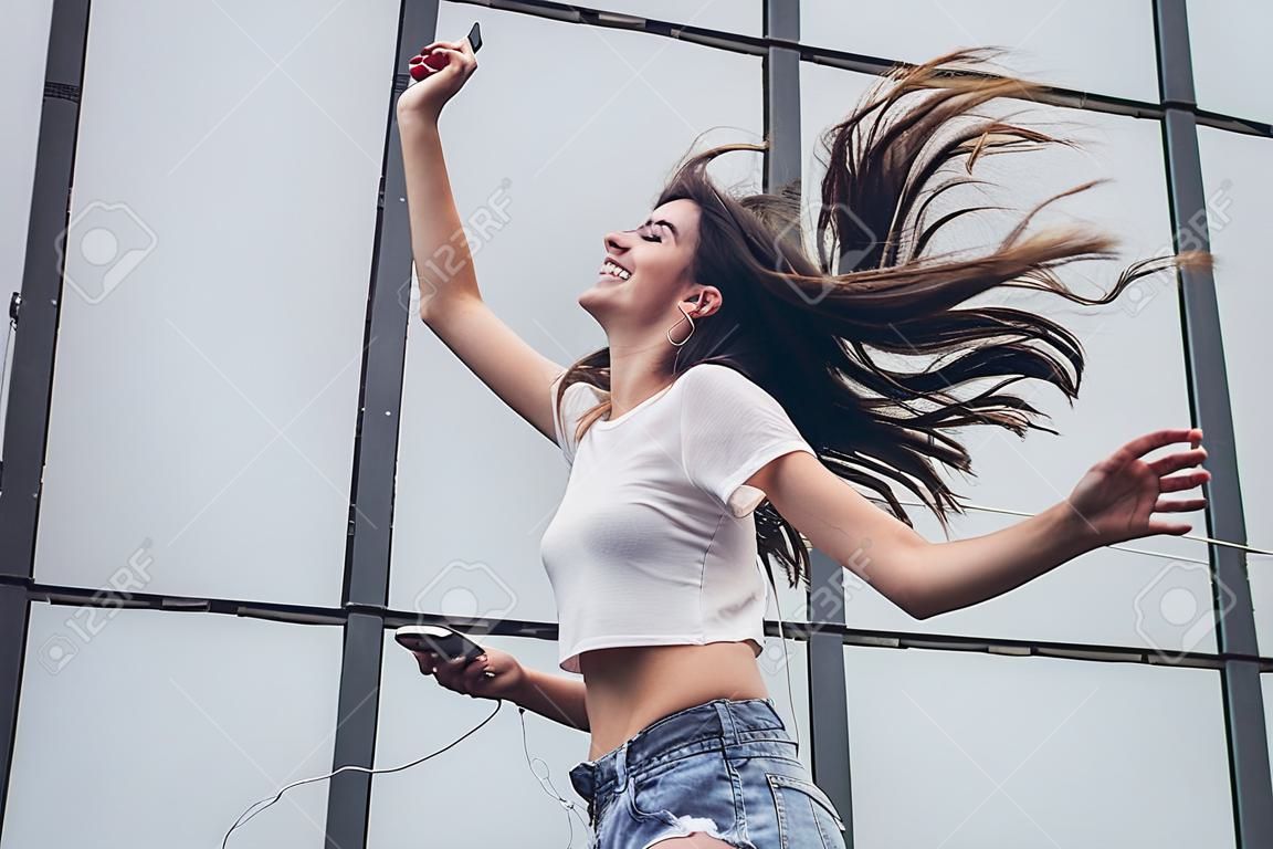 Beautiful smiling teenager with hair flying is having fun outdoors on a gray background. Young woman in earphones with mobile phone in hand is listening to music, smiling and singing.
