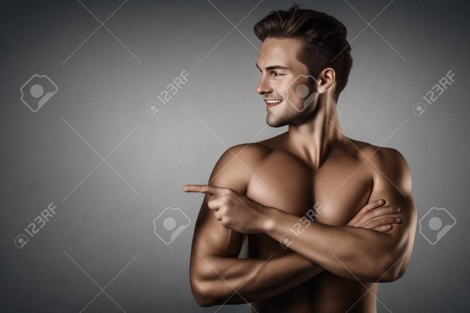 Handsome young man isolated. Portrait of shirtless muscular man is standing on grey background and pointing to the side. Showing product.