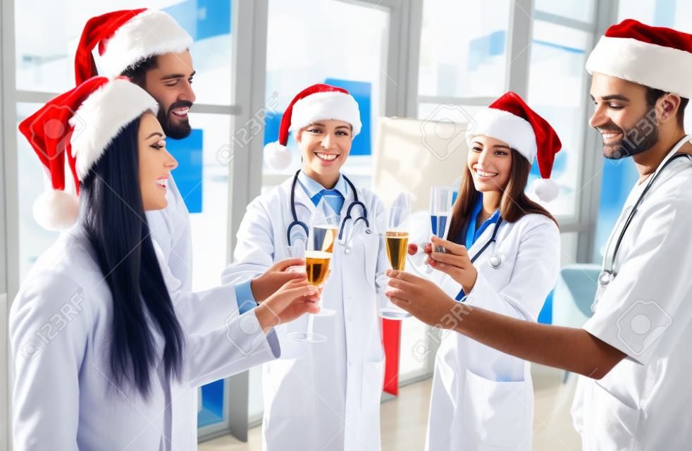 즐거운 성탄절 보내시고 새해 복 많이 받으세요! 직장에서 겨울 방학을 축하하는 의사 그룹. 제복을 입은 의료진과 함께 샴페인을 마시는 산타클로스 모자.