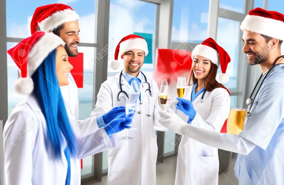 즐거운 성탄절 보내시고 새해 복 많이 받으세요! 직장에서 겨울 방학을 축하하는 의사 그룹. 제복을 입은 의료진과 함께 샴페인을 마시는 산타클로스 모자.