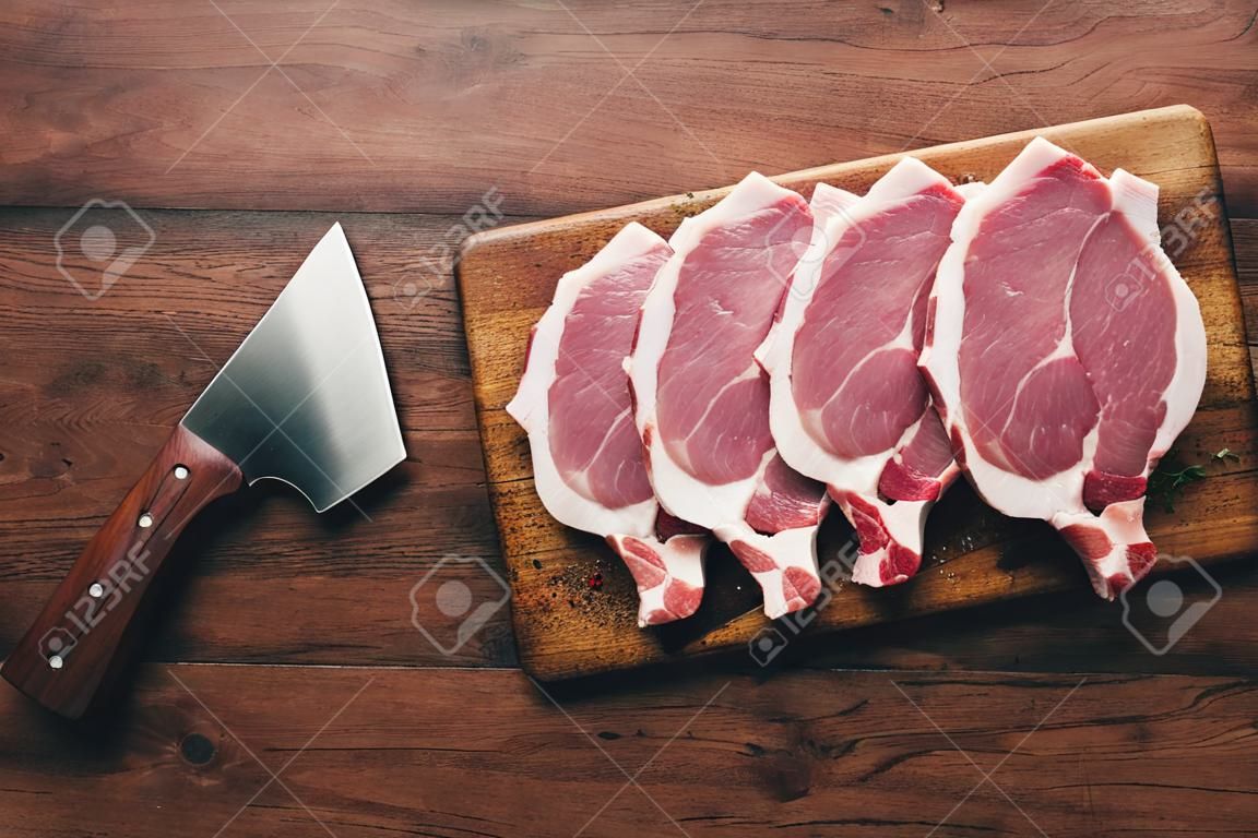 Raw pork chops, meat on rustic wooden cutting board prepared for cooking with garlic, thyme, spices and pepper. Old wood kitchen table, top view