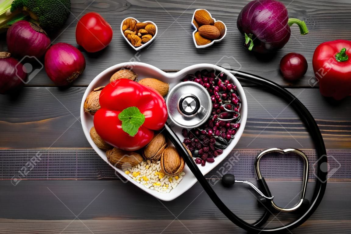 Products good for heart and blood vessels. Vegetables, fruits, nuts in heart shaped bowl near stethoscope on dark wooden background top view