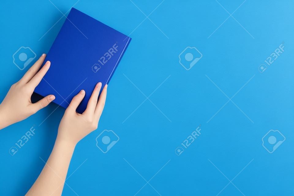 Reading for leasure. Hands take hardback book with empty cover on blue background top view space for text