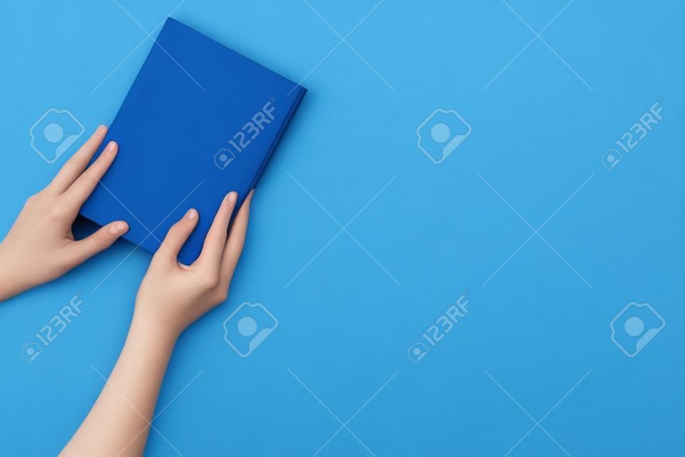 Reading for leasure. Hands take hardback book with empty cover on blue background top view space for text