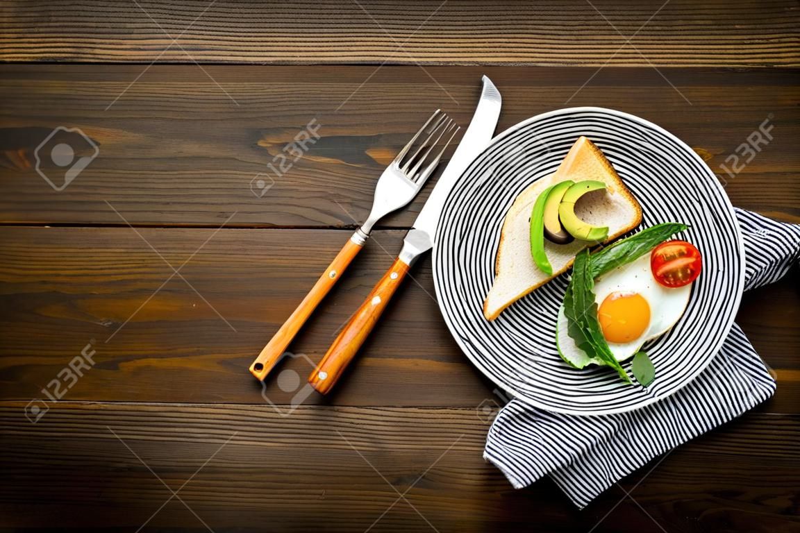 Receita de ovos fritos para o café da manhã vegetariano. Ovos fritos com tomate cereja e vegetação perto de torrada com abacate no espaço de cópia superior de madeira escura