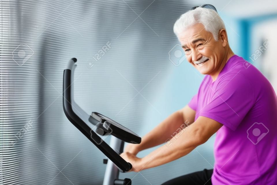 старый старший мужчина езда на велосипеде, упражнения, тренировки в тренажерном зале с современным велотренажером