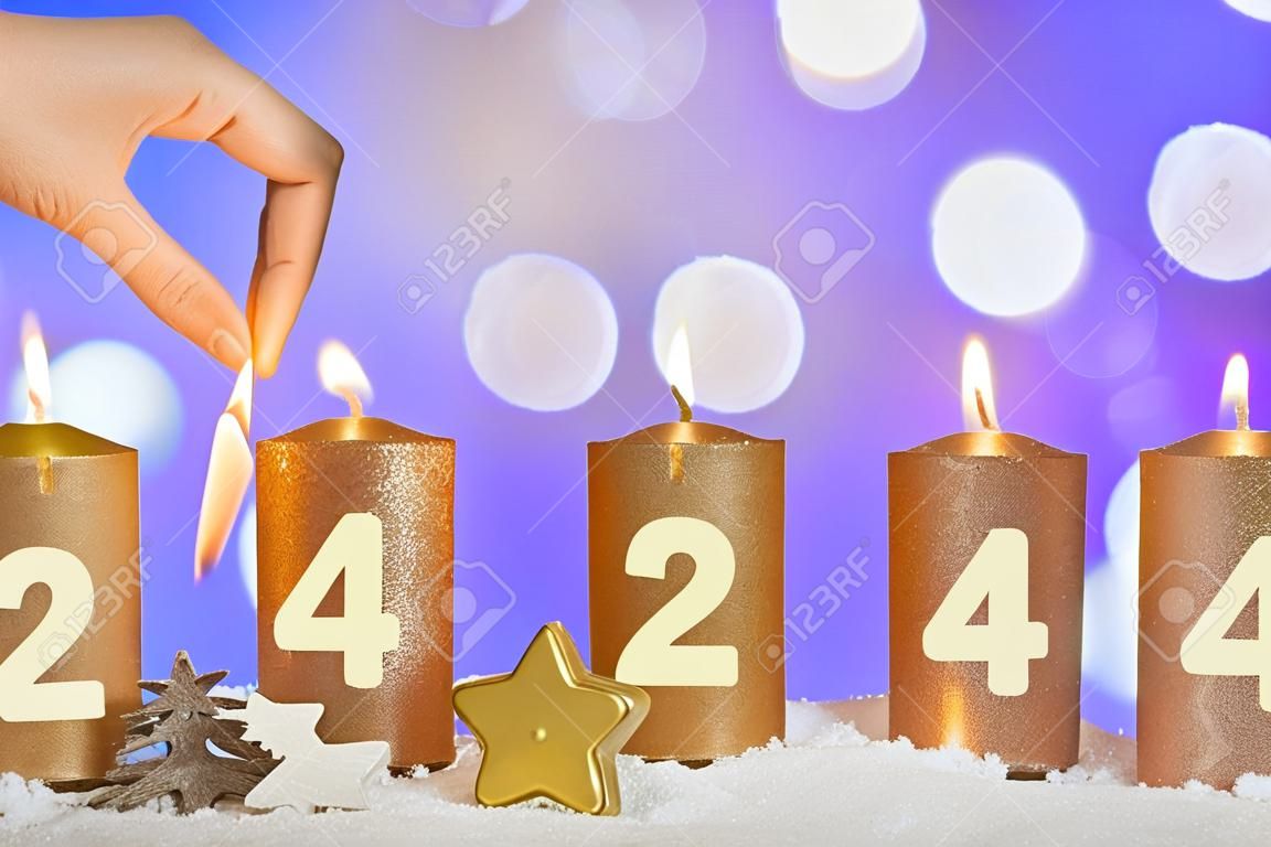 Four numbered gold advent candles with first candle lit by hand with match and christmas decoration lying in snow as template