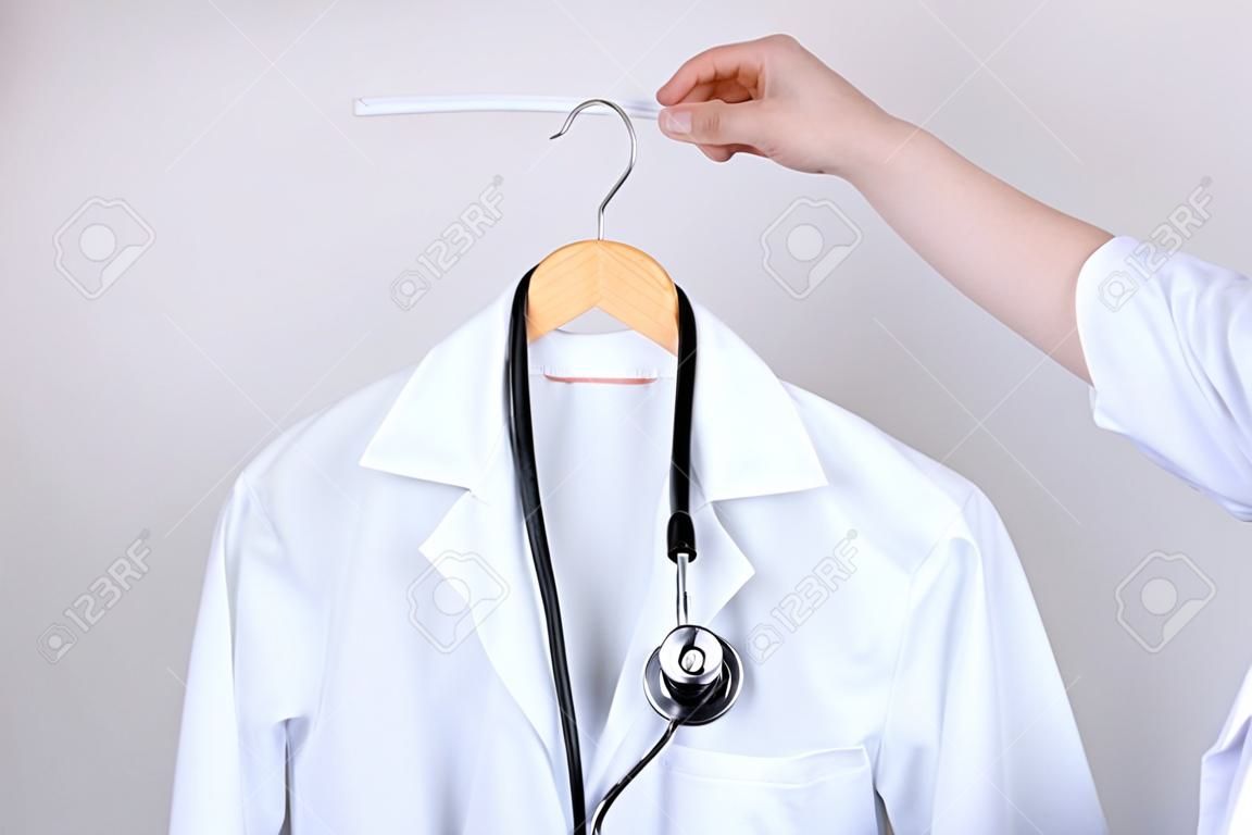 Koncepcja wolnych miejsc pracy. przycięte z bliska zdjęcie dłoni trzymającej biały fartuch lekarza na wieszaku odizolowanym na szarym tle