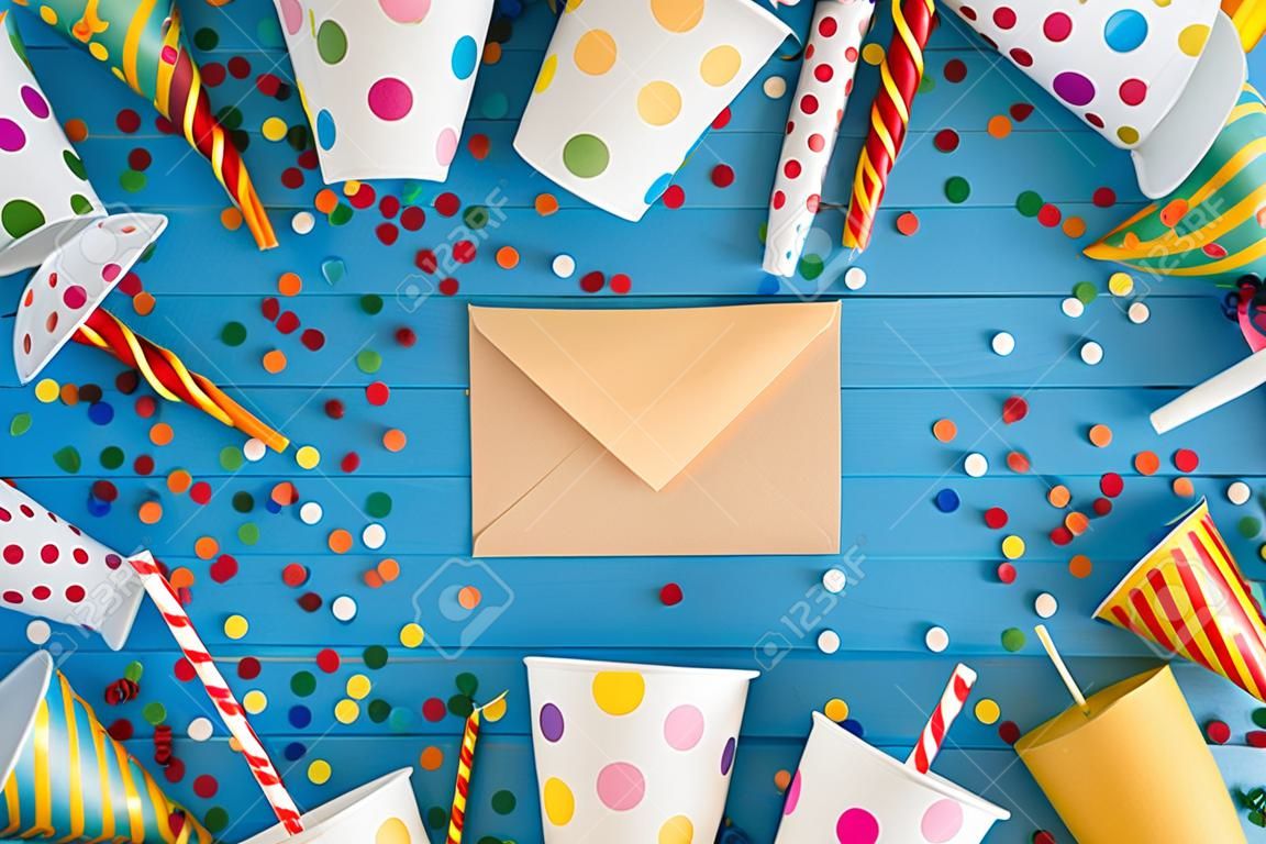 Top view photo of birthday party composition closed craft paper envelope in the middle spiral candles pipes straws hats balloons confetti paper cups and plates on isolated blue wooden table background