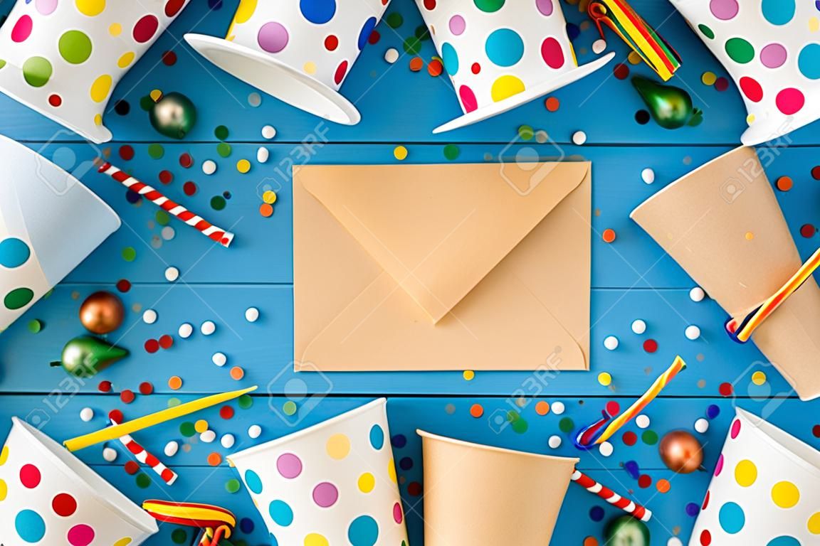 Top view photo of birthday party composition closed craft paper envelope in the middle spiral candles pipes straws hats balloons confetti paper cups and plates on isolated blue wooden table background