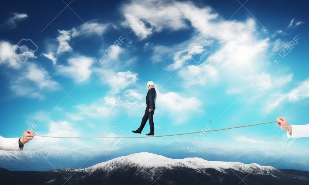 Biznesmen chodzący z zasłoniętymi oczami na linie nad wysokimi górami jako symbol ukrytych zagrożeń i wsparcia. widok nieba i przyrody na tle.
