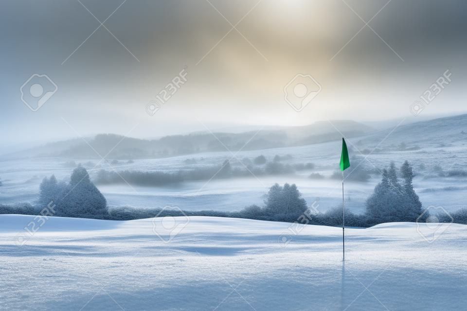 Ein Golfplatz in Schottland an einem schneereichen Wintermorgen im Dezember. Blick von einer grünen mit einer blauen Flagge, mit Bäumen, Nebel und Berge im Hintergrund.