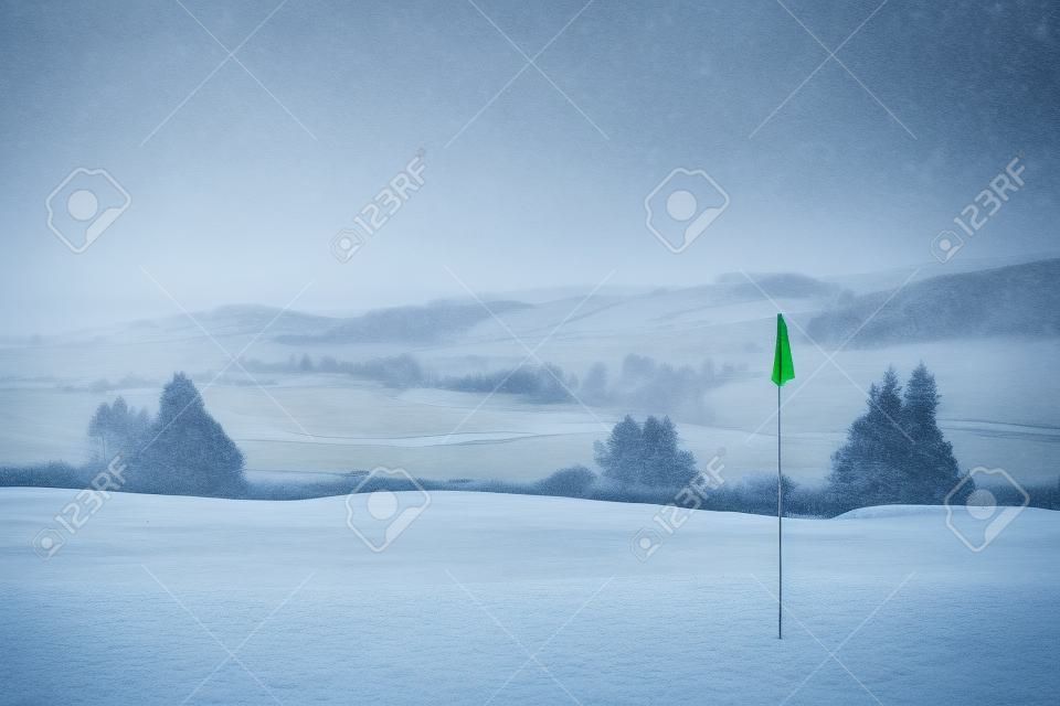 Ein Golfplatz in Schottland an einem schneereichen Wintermorgen im Dezember. Blick von einer grünen mit einer blauen Flagge, mit Bäumen, Nebel und Berge im Hintergrund.