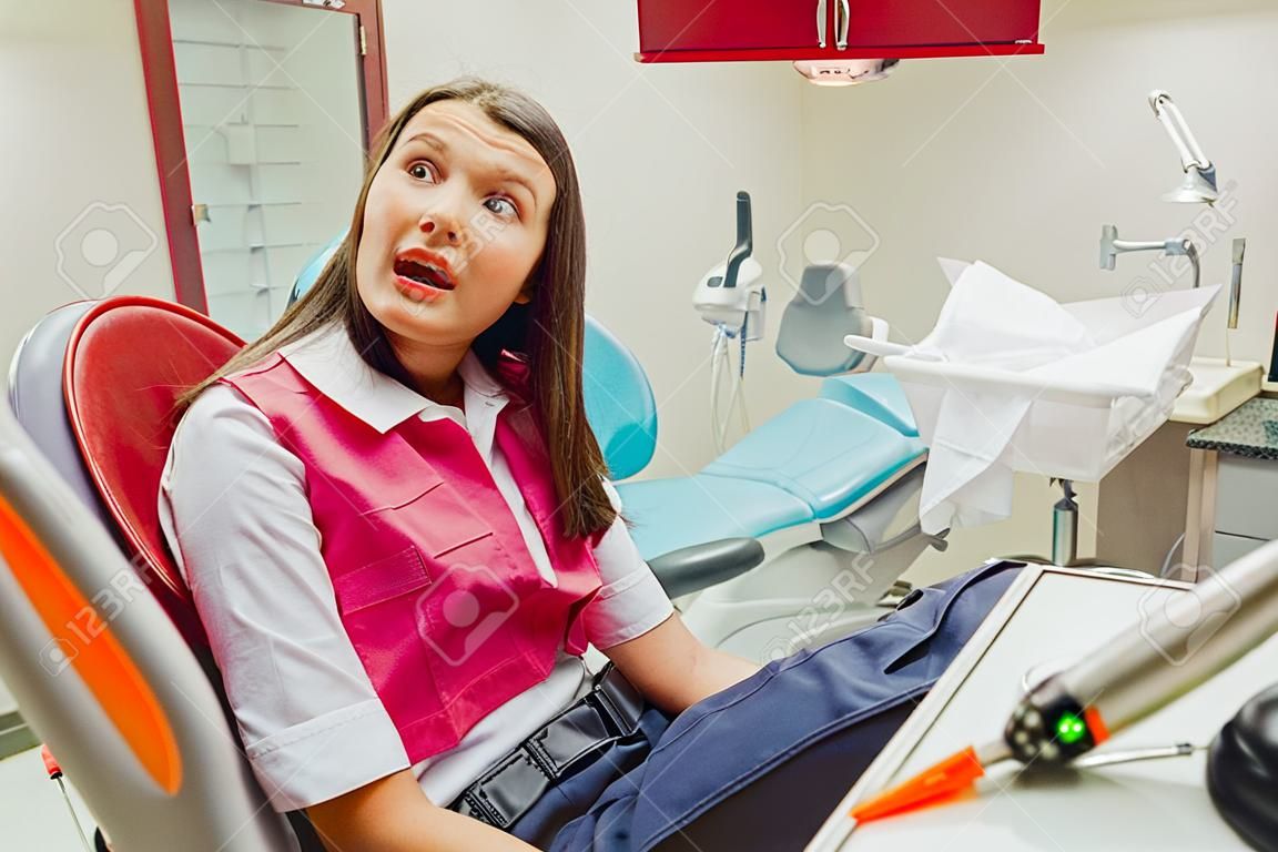 Ritratto del primo piano di una giovane ragazza triste con un dente doloroso, un medico in sedie da ufficio, una clinica di fondo dell'ufficio del dentista isolato. E