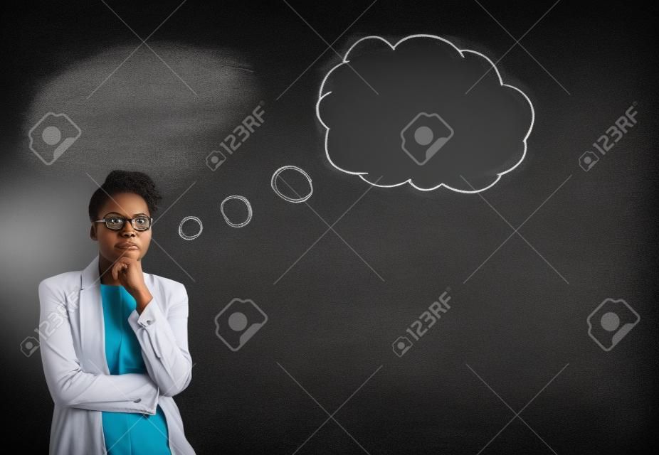 Sud Africa o donna afro-americana nera insegnante o studente con la sua mano sul suo mento, mentre il pensiero nuvola pensiero o bolle in piedi su uno sfondo di gesso lavagna dentro