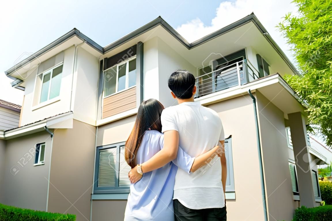 Tylny portret azjatyckiej młodej pary stojącej i przytulającej się, wyglądającej na szczęśliwą przed swoim nowym domem, aby rozpocząć nowe życie. koncepcja rodziny, wieku, domu, nieruchomości i ludzi.