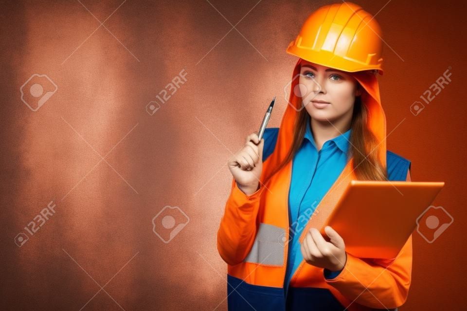 Woman construction worker builder structural engineerin in orange vest red hard helmet holds pen file pad. Safety in industrial work. Studio shot