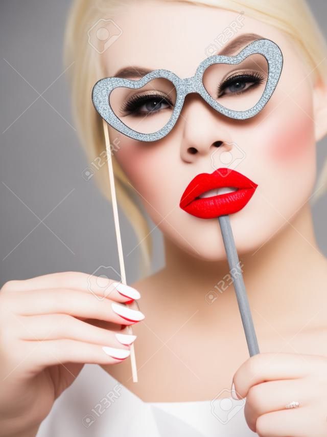 Happy blonde woman holding carnival accessories on stick fake red lips and paper heart shaped glasses, having fun. On grey wall background