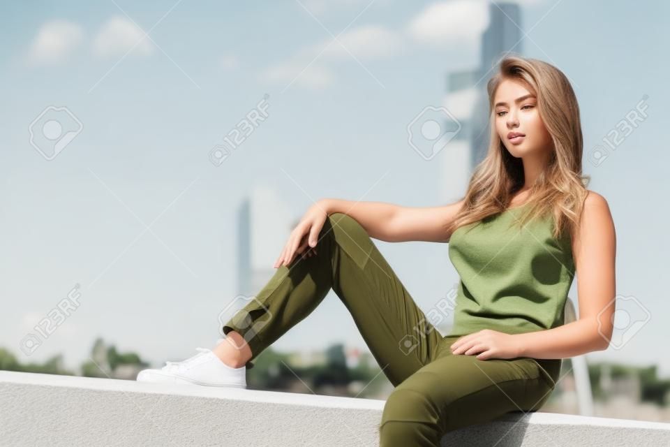 Giovane bella modella donna seduta sul muro di cemento che indossa canotta bianca e pantaloni verde oliva. Femmina che cammina all'aperto durante il caldo clima estivo.