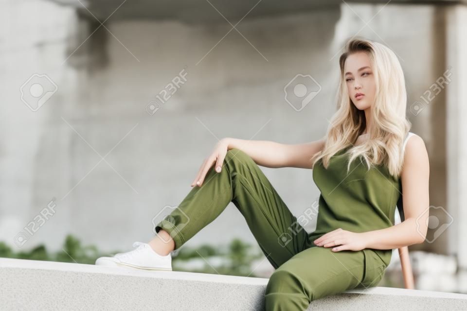 Young pretty fashion model woman sitting on concrete wall wearing white tank top and olive green trousers. Female walking outdoor during warm summer weather.