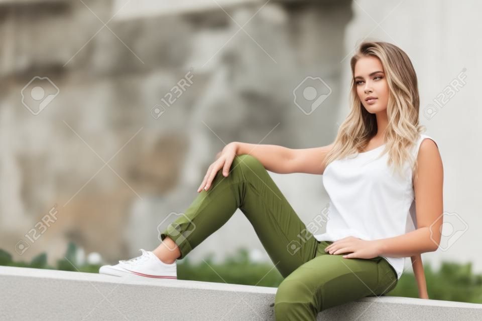 Young pretty fashion model woman sitting on concrete wall wearing white tank top and olive green trousers. Female walking outdoor during warm summer weather.