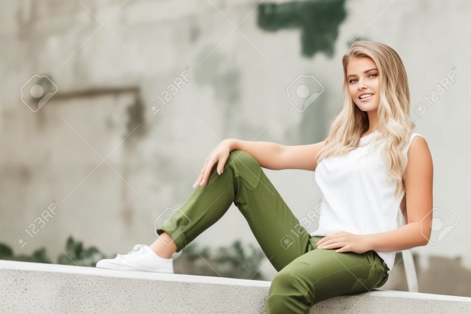 Junge hübsche Mode-Modell-Frau sitzt auf Betonwand mit weißem Trägershirt und olivgrüner Hose. Weibliche Spaziergänge im Freien bei warmem Sommerwetter.