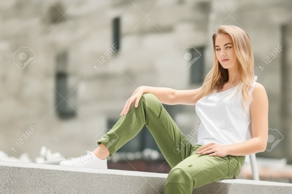 흰색 탱크 탑과 올리브 녹색 바지를 입고 콘크리트 벽에 앉아 젊은 예쁜 패션 모델 여자. 따뜻한 여름 날씨에 야외에서 걷는 여성.