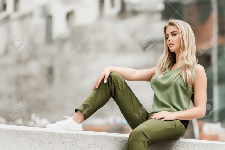 Mujer bonita joven modelo sentada en la pared de hormigón con camiseta blanca y pantalón verde oliva. Mujer caminando al aire libre durante el clima cálido de verano.