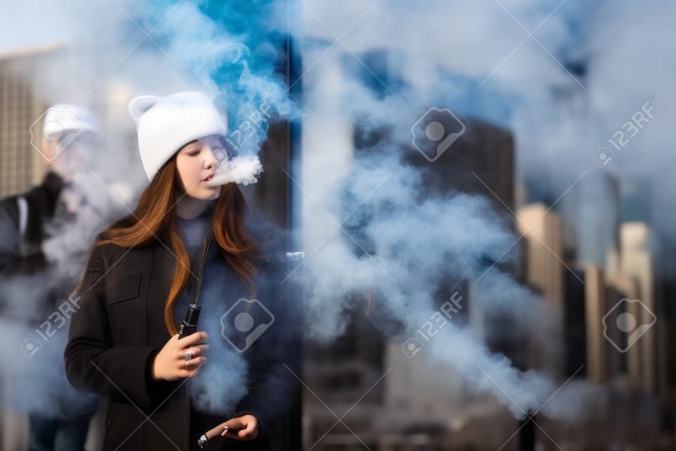Vape teenager. Young pretty white girl in checkered coat and black cap smoking an electronic cigarette opposite glass building on the street in the spring. Bad habit. Vaping activity.