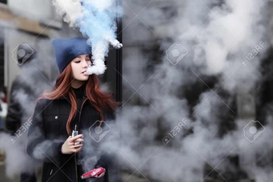 Vape teenager. Young pretty white girl in checkered coat and black cap smoking an electronic cigarette opposite glass building on the street in the spring. Bad habit. Vaping activity.