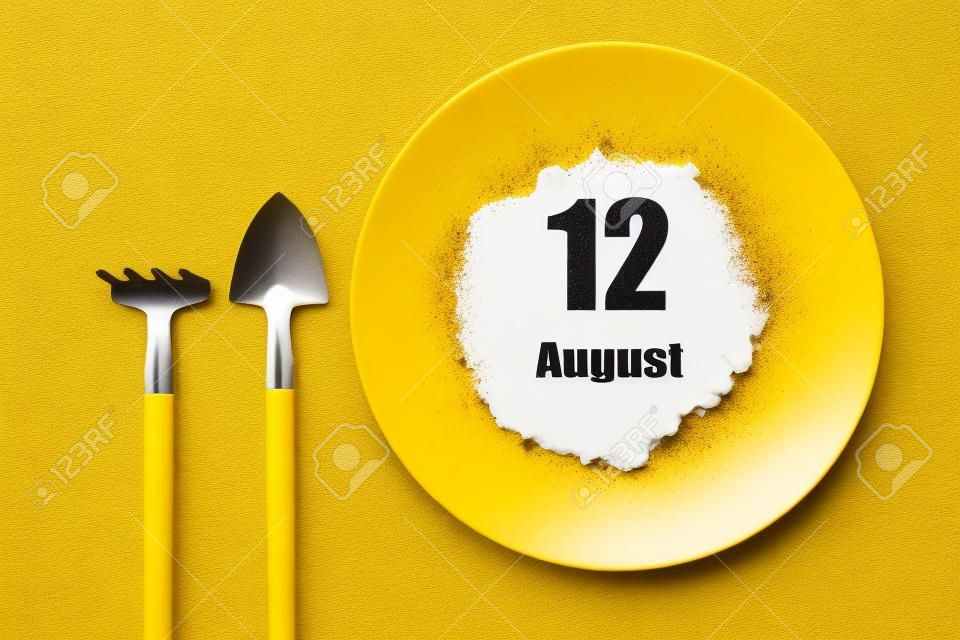 August 12nd. Day 12 of month, Calendar date. White plate of soil with a small spatula and rake on yellow background. Summer month, day of the year concept