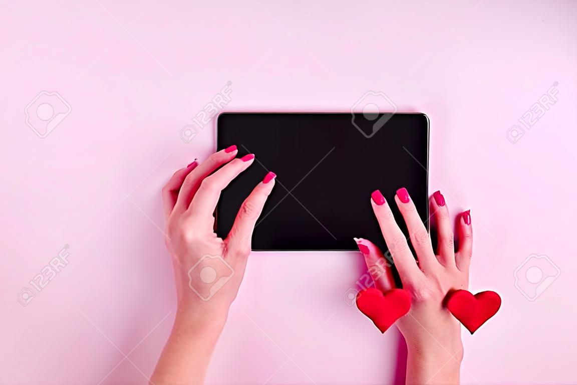 Online shopping concept. Female hands on a black blank tablet screen with red heart shape on a pink background, copy space, minimalism, flat lay. Valentine day concept
