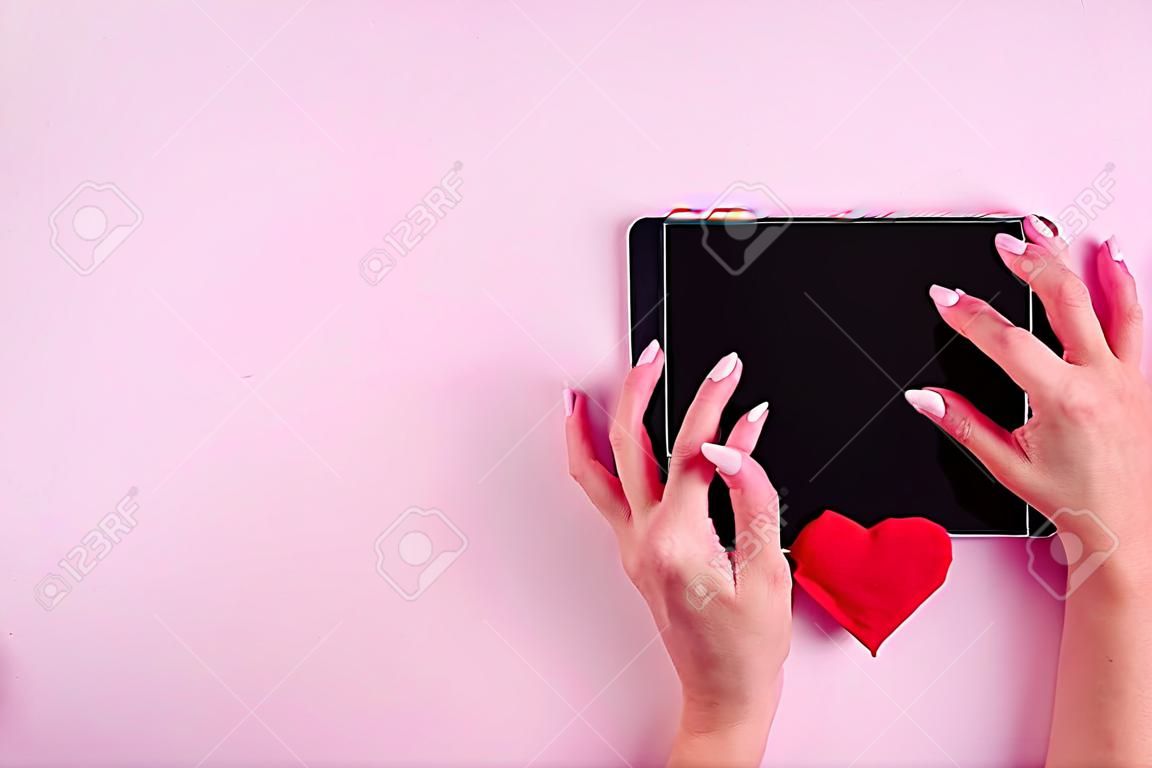 Online shopping concept. Female hands on a black blank tablet screen with red heart shape on a pink background, copy space, minimalism, flat lay. Valentine day concept