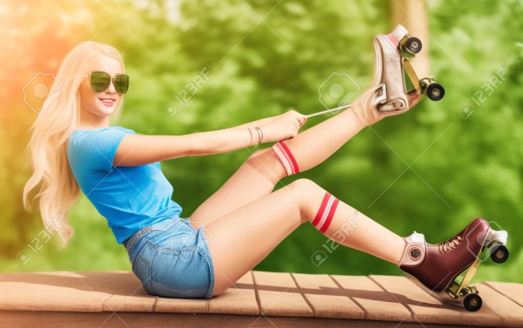 Ritratto di una bella ragazza bionda stringere i lacci seduto su una panchina in un vintage pattini a rotelle, indossando pantaloncini, golf e una T-shirt. Guardando la fotocamera Caldo giorno d'estate All'aperto.