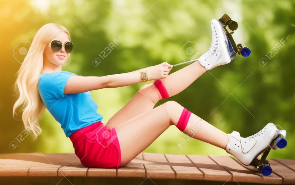 Retrato de una hermosa joven rubia apretar los cordones que se sientan en un banco en un patines de época, con pantalones cortos, campos de golf y una camiseta. Mirando a la cámara. Día caluroso de verano. Al aire libre.