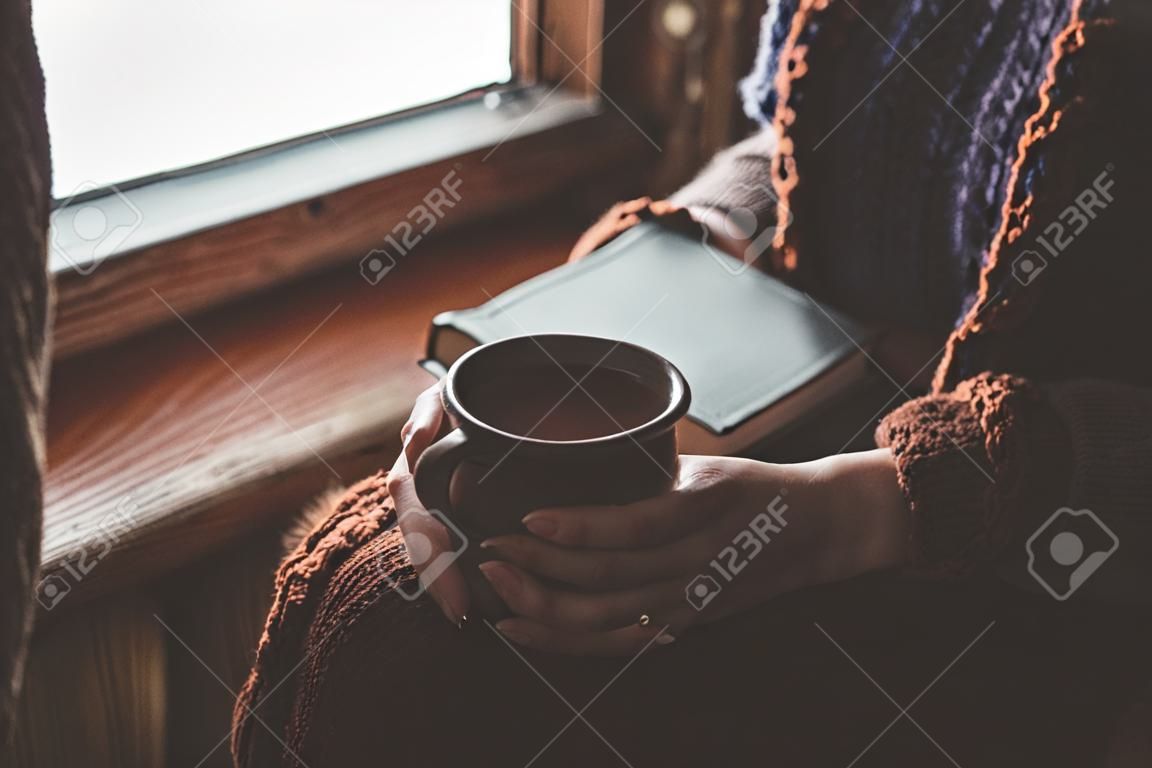 Meisje in warme wollen trui zitten op vensterbank, thee drinken en een boek lezen. Winter weekends in oud log house. Koud besneeuwd weer. Gezellig huiselijk concept.