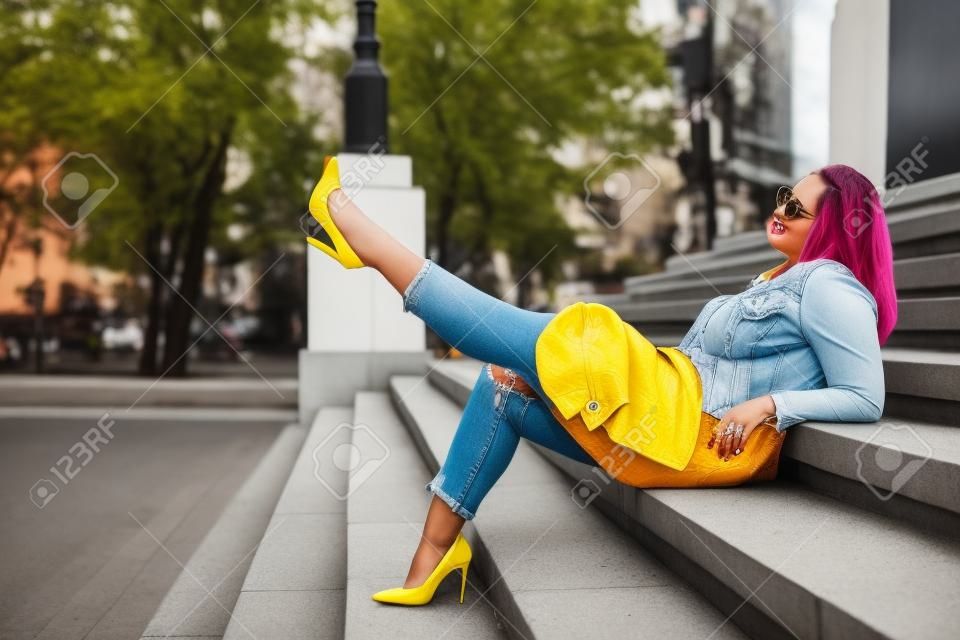 Preriry穿著明亮的五顏六色的夾克的年輕女子，撕開牛仔褲和腳跟鞋走在城市街道上。休閒時尚，加上尺碼模特兒。