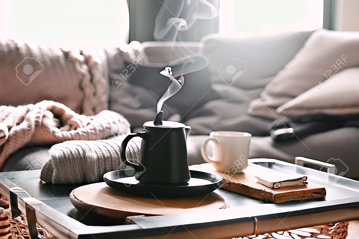 Натюрморты в домашнем интерьере гостиной. Свитера и чашка чая с паром на сервировочном подносе на кофейном столике. Завтрак на диване в утреннем солнечном свете. Уютная осенняя или зимняя концепция.