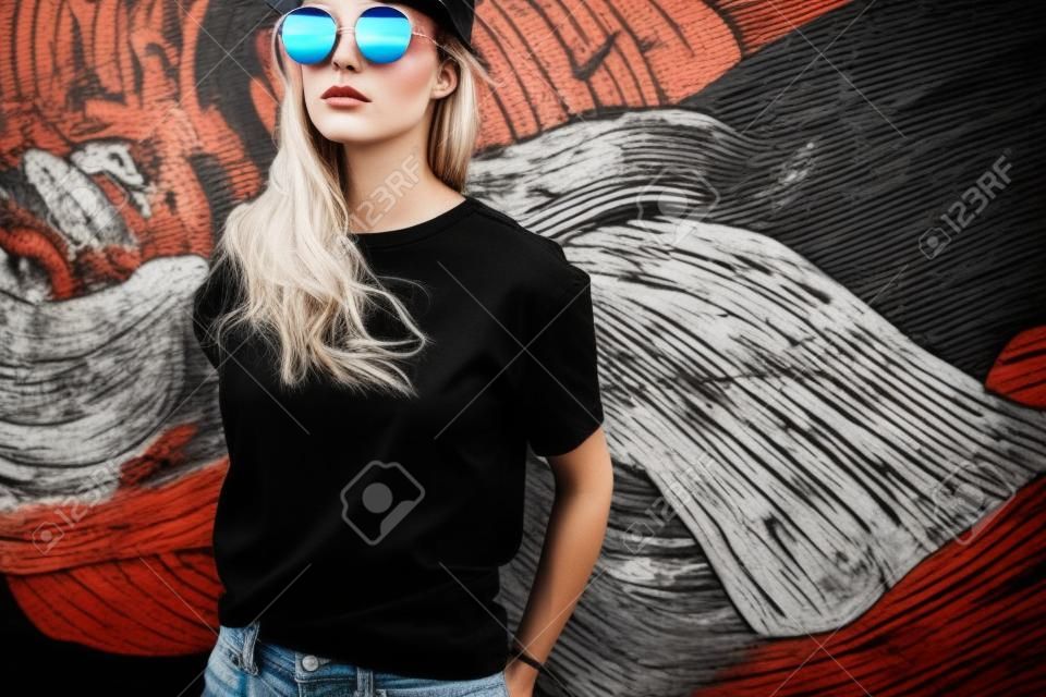 Model wearing plain black t-shirt and hipster sunglasses posing against street wall, teen urban clothing style, mockup for tshirt print store