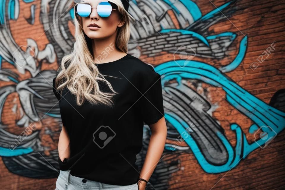 Model wearing plain black t-shirt and hipster sunglasses posing against street wall, teen urban clothing style, mockup for tshirt print store