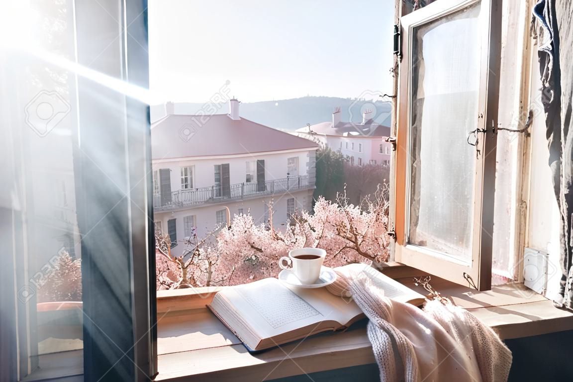 Stare okno z widokiem od środka na kwitnące drzewo. Otwarta książka, filiżanka herbaty lub kawy, okulary i sweter z dzianiny na parapecie. Koncepcja przytulny weekend wiosna.