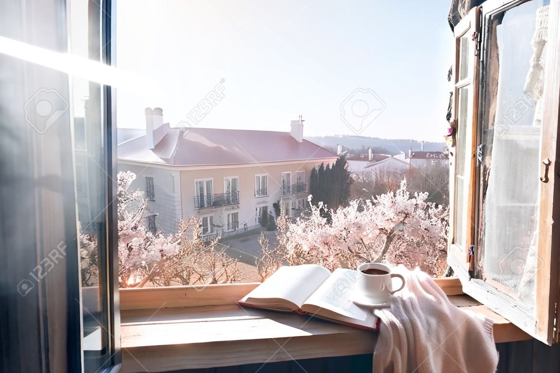 Old window with the view from inside into blooming tree. Opened book, cup of tea or coffee, glasses and knitted sweater on windowsill. Cozy spring weekend concept.