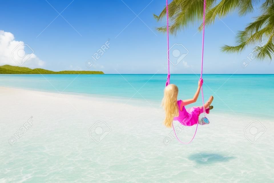 Il punto di vista posteriore della ragazza con il rosa sente divertirsi sull'oscillazione che appende sull'albero alla spiaggia tropicale con sabbia bianca. Vacanza di lusso sull'isola paradisiaca.